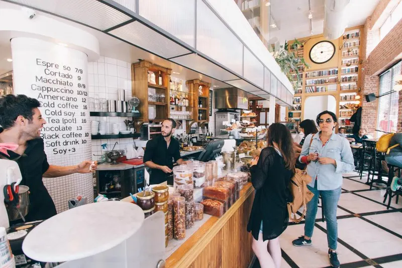 Kafe ramai dengan papan menu minuman, barista di balik meja, dan pelanggan yang menunggu pesanan
